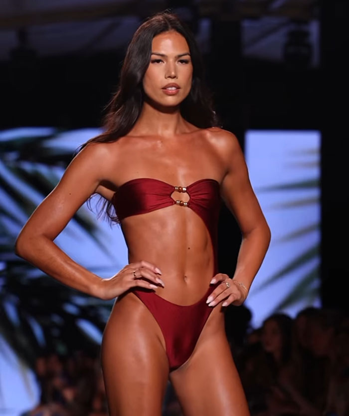 Trans model confidently walking runway in a maroon bikini at Miami Swim Week, showcasing bold and stylish beachwear.