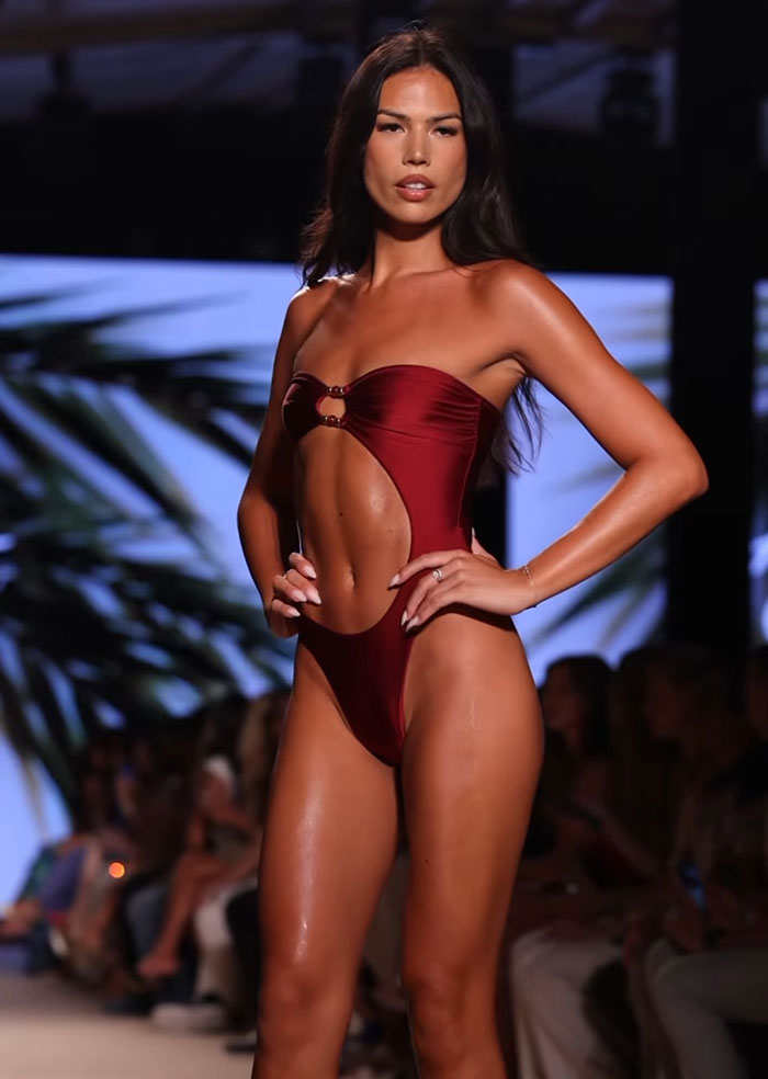 Trans model confidently walking runway in burgundy bikini at Miami Swim Week with massive support from the crowd.