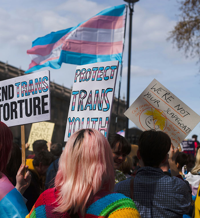 Protesters at rally holding signs supporting trans youth rights with a transgender pride flag waving in the background.