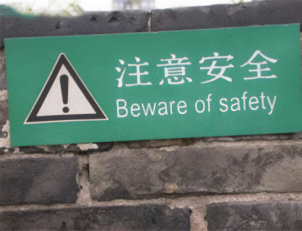Green warning sign with confusing English translation Beware of safety and Chinese characters on a brick wall.