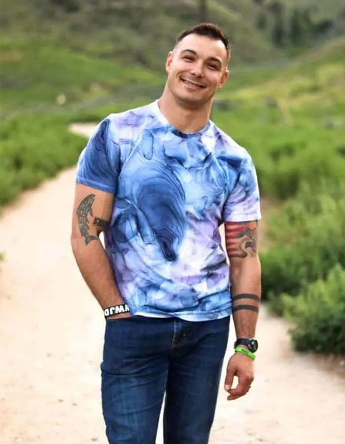 Man with tattoos wearing a blue patterned shirt standing on a dirt path with greenery, related to tragic cause of passing.