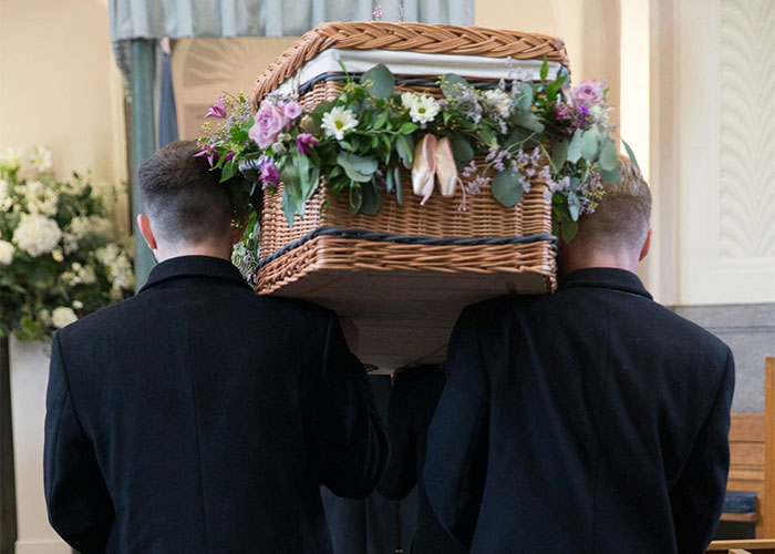 Two men in black suits carrying a flower-adorned wicker casket during a funeral service, sharing unbelievable experiences.