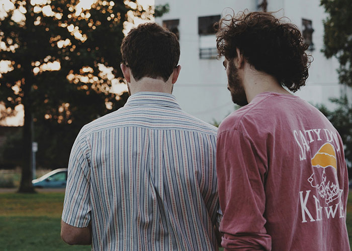 Two men sharing their most unbelievable experiences, standing outdoors near trees and a building at sunset.