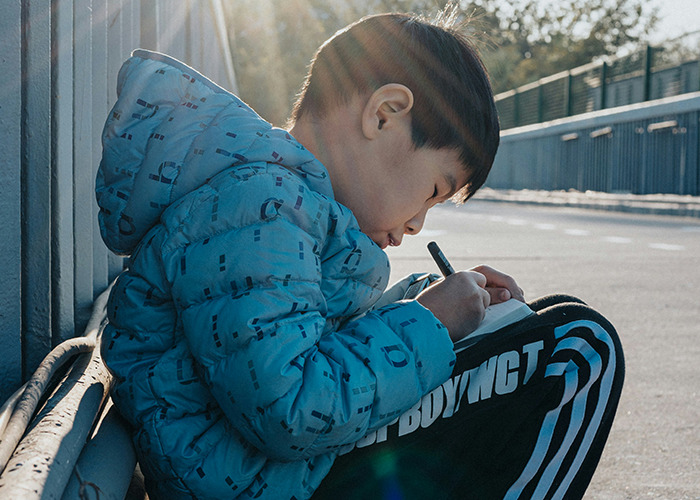 Young boy in a blue jacket intensely writing down his unbelievable experiences outdoors on a sunny day.