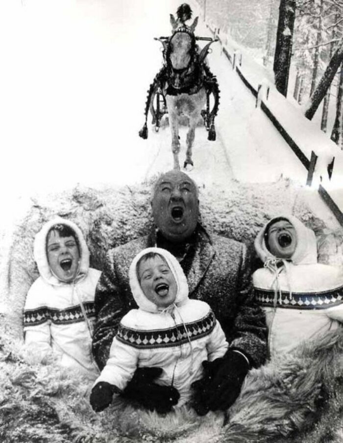 Black and white historical photo of a man and three children sledding in the snow with a horse-drawn sleigh behind them