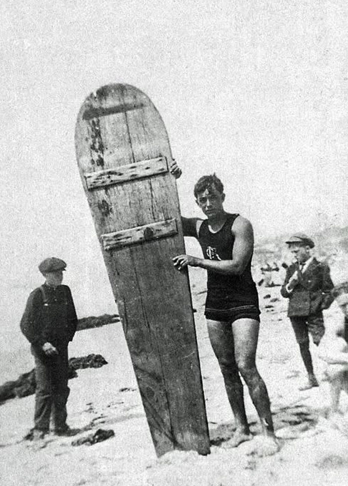 Young man on a beach holding a large wooden surfboard, captured in a rare historical photo from the early 20th century.