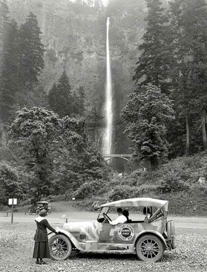 Rare historical photo of early 20th century car and people near a tall waterfall surrounded by dense forest.