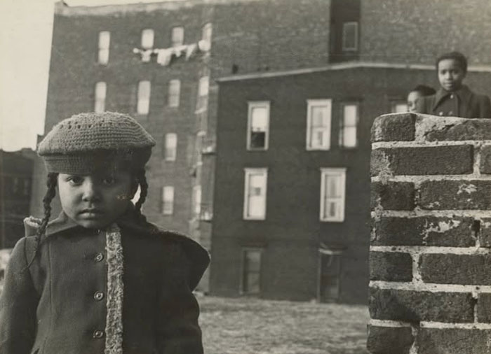 Rare historical photo of a young girl in vintage clothing standing outdoors in an urban neighborhood with brick buildings.