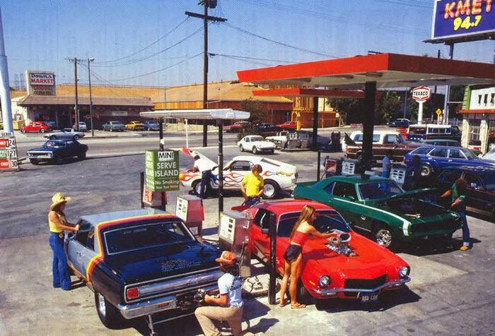 Rare historical photo of a vintage gas station with classic cars and people filling up in a busy urban setting.