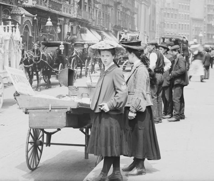 Two women wearing vintage clothing and hats beside a street vendor cart in a rare historical photo from the early 1900s.