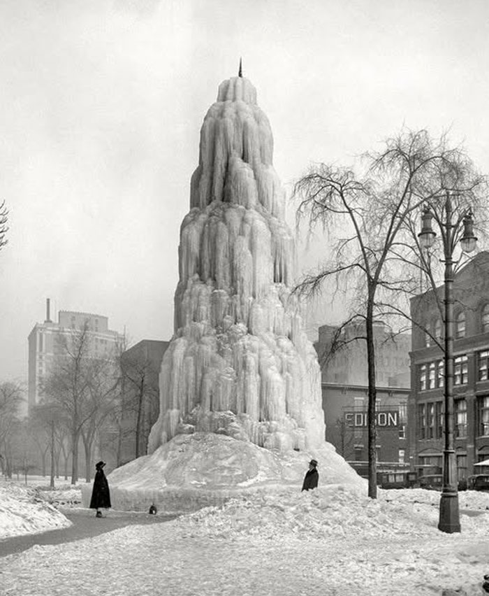 Rare historical photo of a large frozen ice tower in a city park with two people standing nearby in winter.