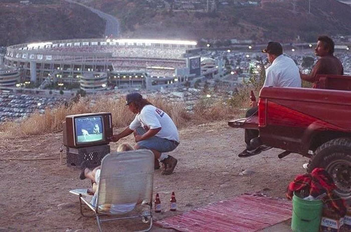 Rare historical photo of people watching a TV baseball game outdoors near a stadium at dusk in the 1980s.