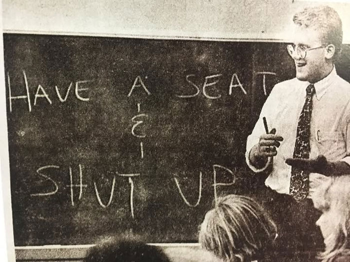 Black and white rare historical photo of a teacher smiling while students sit in front of a chalkboard with sarcastic writing.