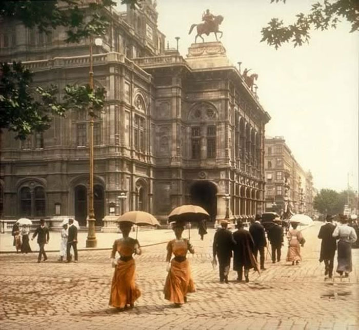 Rare historical photo of early 20th century pedestrians with parasols walking near a grand European building on a cobblestone street.