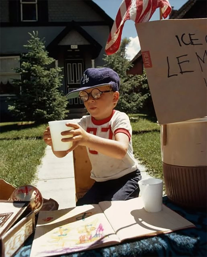 Young boy wearing glasses and a baseball cap selling lemonade at a stand in a rare historical photo outdoors.