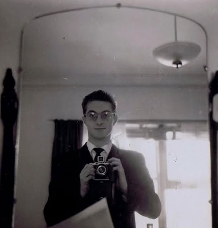 Young man wearing glasses and suit taking a self-portrait with a vintage camera in a rare historical photo.