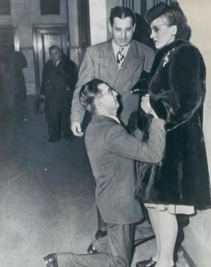 Rare historical photo showing a man kneeling and proposing to a woman dressed in vintage clothing, with another man watching.