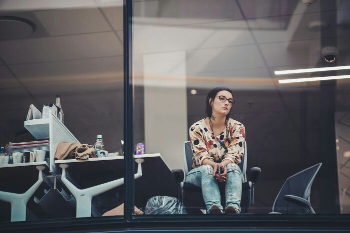 Young woman sitting alone in office behind glass, reflecting on ethical and unethical life hacks to win in the real world.