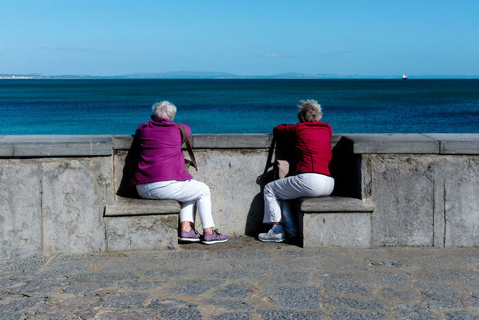 Two elderly women sitting on a stone bench by the sea, captured in stunning street photos that show candid moments.