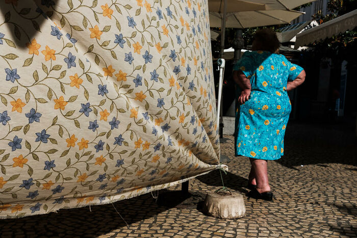 Colorful street photo of a woman in a blue floral dress walking past a patterned fabric on a sunny cobblestone street.