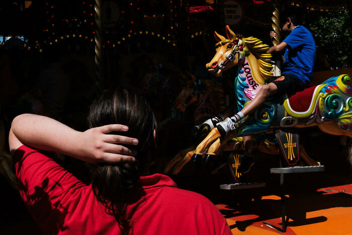 Child in red shirt watching colorful carousel horse with boy riding during vibrant street photo moment