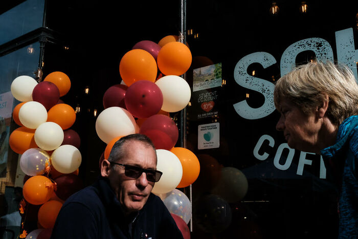 Man wearing sunglasses in a candid street photo with colorful balloons outside a coffee shop captured by Polly Rusyn.
