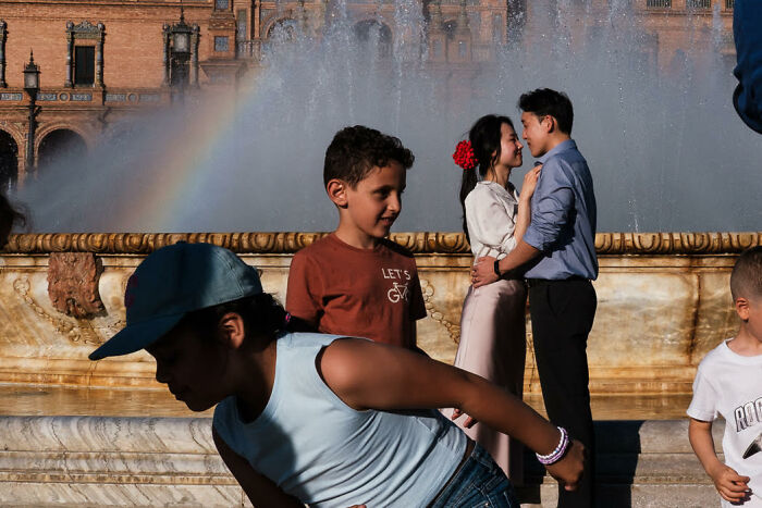 Children playing near a fountain while a couple shares a candid moment in colorful, stunning street photography.