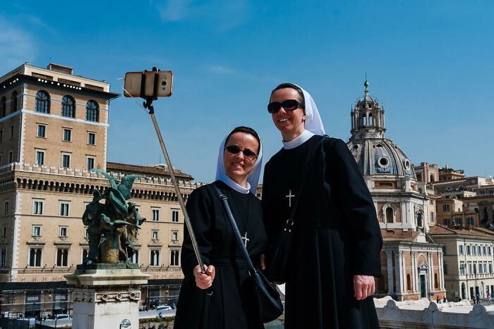 Two nuns taking a colorful candid street photo with a selfie stick in front of historic city buildings on a sunny day.