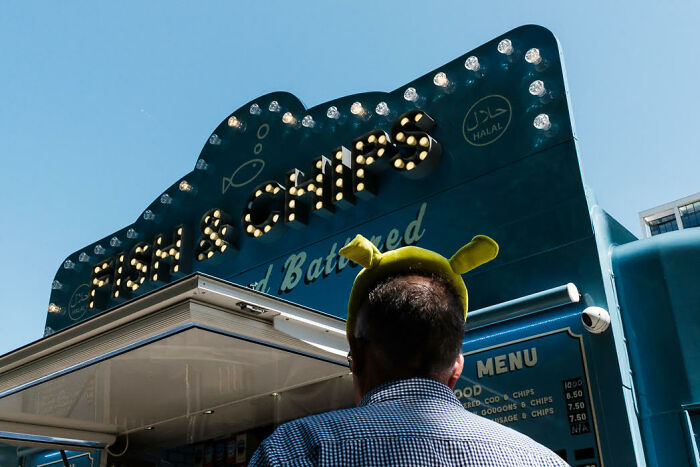 Man wearing Shrek ears standing in front of a colorful fish and chips stand in a candid street photo.
