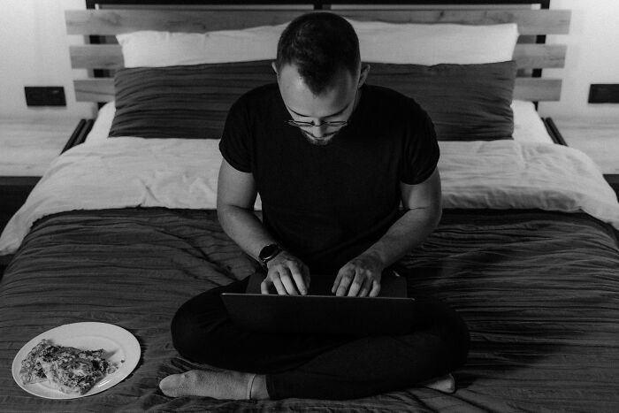 Man sitting cross-legged on a bed, focused on typing on a laptop with a slice of pizza on a plate nearby in an unpopular opinions poll.