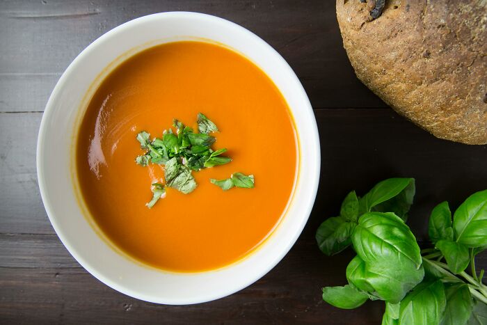 Bowl of creamy orange soup garnished with herbs next to fresh basil and a rustic whole grain bread on dark wood table.