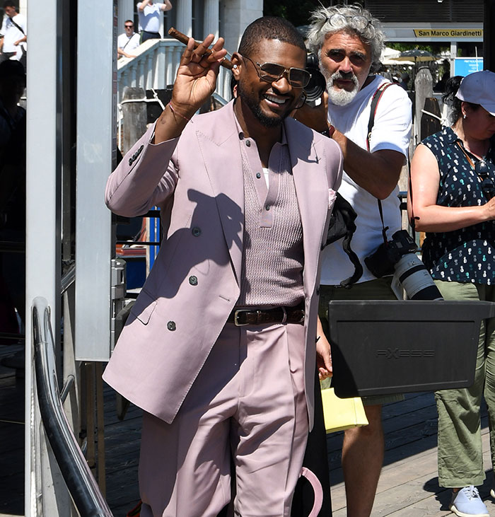 Man in a pink suit smiling and waving outdoors with photographers behind, related to Charlize Theron and Bezos wedding guests topic