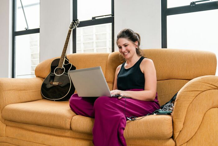 Young woman smiling on a couch with a laptop and a guitar nearby, focusing on epicly unhinged ways to get back at cheating exes.