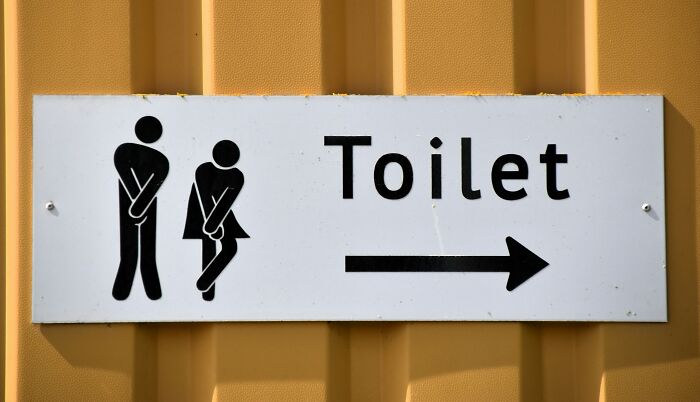 Sign indicating toilet direction with icons of a man and woman holding their lower bodies, illustrating casual habits that may be damaging.