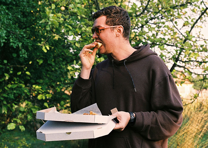 Man in a dark hoodie eating pizza outdoors with greenery in the background, captured in a casual moment on security tape.
