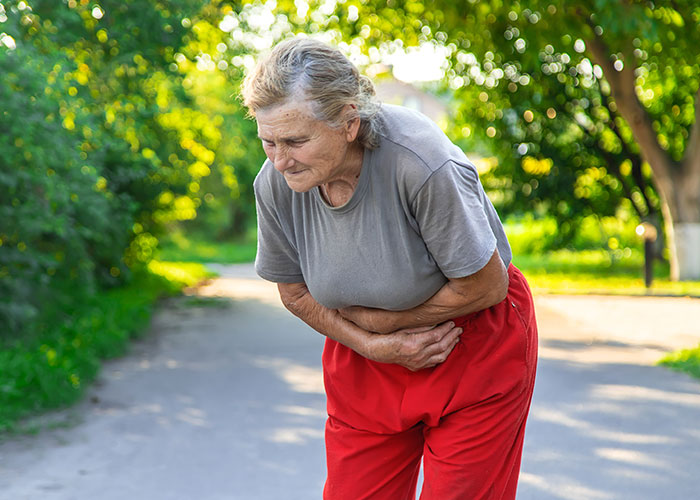 Elderly woman outdoors clutching stomach in discomfort, a random moment possibly caught on security tape.