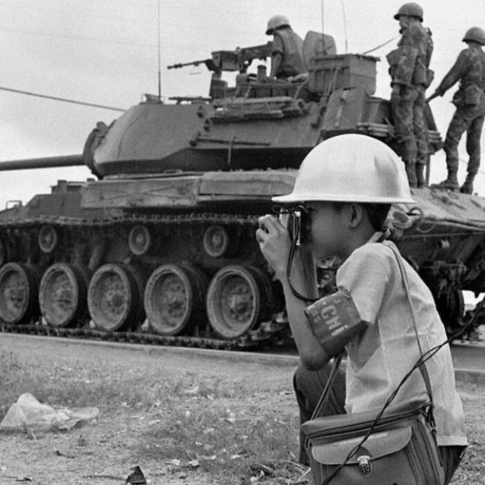 Child wearing helmet and armband using a camera near a military tank with soldiers, illustrating weird things about history.