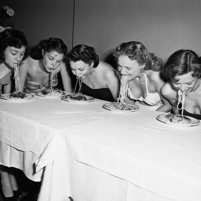Five women in vintage dresses eating spaghetti without utensils at a table, illustrating weird things about history.