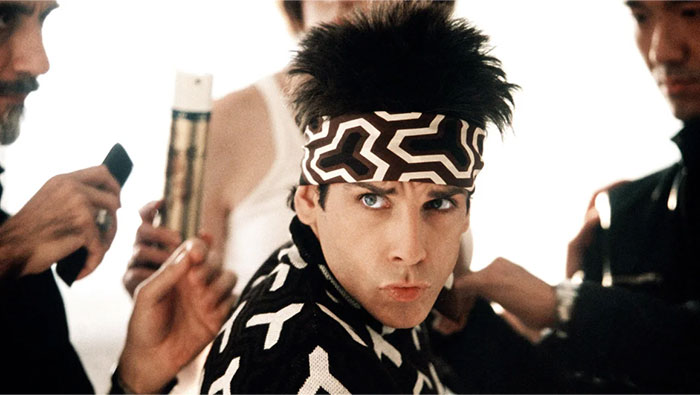 Actor wearing a patterned headband and jacket, surrounded by stylists preparing him for a photo shoot on set.