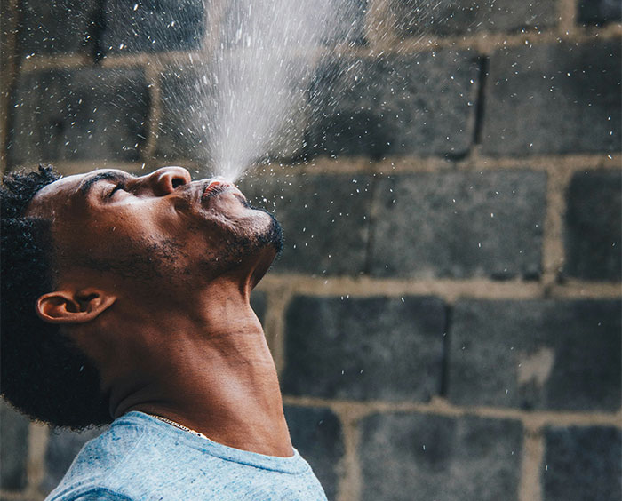 Man in blue shirt looking up while spitting water droplets against a textured stone wall background.