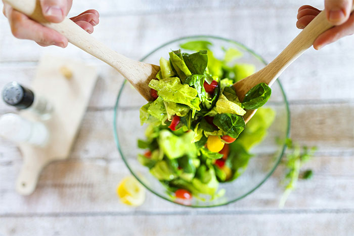 Fresh salad being tossed in a glass bowl using wooden spoons, highlighting healthy food and lifestyle concepts.