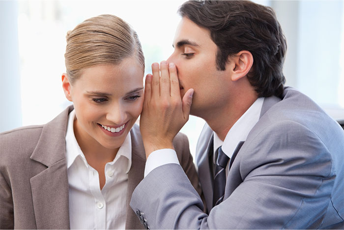 Man whispering a secret to a smiling woman in business attire, illustrating relationship icks shared by women.