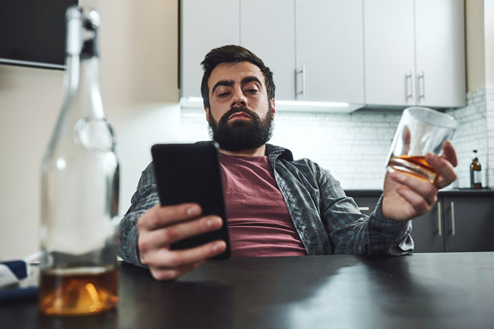 Man with a beard holding a glass of alcohol and looking at his phone in a kitchen, depicting mentally unstable boyfriend scene.