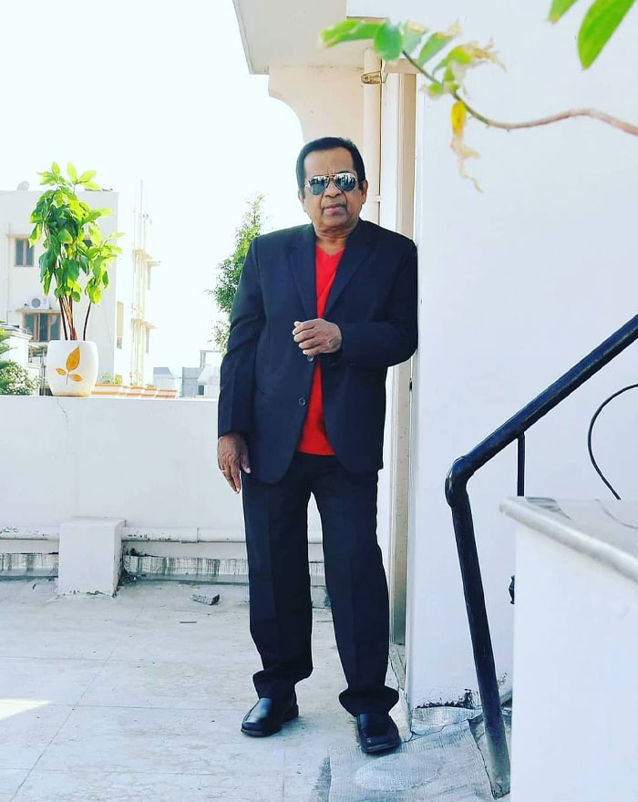 Actor Brahmanandam Kanneganti standing outdoors on a rooftop, wearing a black suit and sunglasses, representing actors in the most movies across genres.