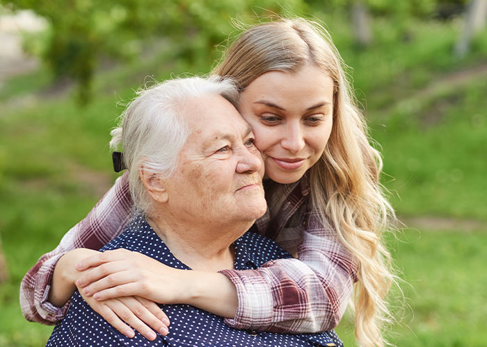 Young woman warmly hugging an elderly woman outdoors, capturing a wholesome interaction between strangers in a green setting.