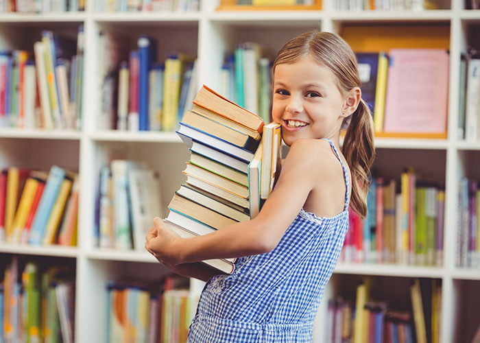 Young girl smiling and holding a large stack of books in a library, representing wholesome interactions with strangers.