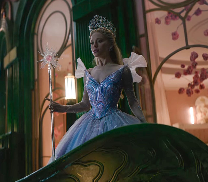 Ariana Grande in a blue gown and tiara holding a wand, showcasing an entirely visible ribcage in a cinematic scene.