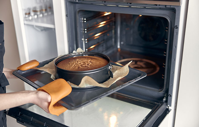 Person wearing oven mitts placing a cake in a pan lined with parchment paper into an oven, highlighting product design flaws.