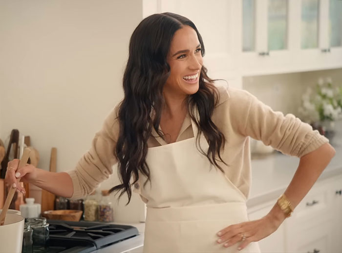 Meghan Markle smiling in a kitchen wearing an apron while sharing her kitchen hack in a bright home setting.