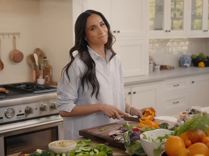 Meghan Markle in kitchen sharing cooking hack, preparing vegetables on wooden cutting board with fresh produce nearby
